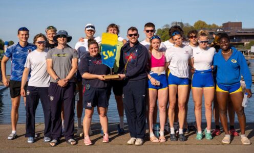 A photo of South West GB Performance Development Academy rowers and coaches, standing next to the water at the National Watersports Centre in Nottingham, holding a trophy painted blue and gold, bearing the letters SW, after their overall win at the 2025 British Rowing Olympic Pathway Regatta